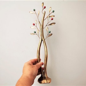 May include: A gold-colored metal decorative tree sculpture with leaves and berries in red, green, and gold. The sculpture is held by a hand against a white background. The tree has a unique, artistic design.