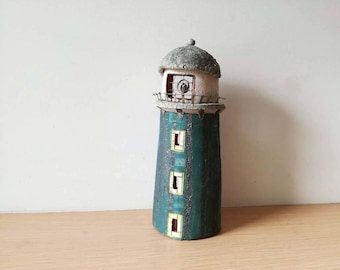 May include: A small, hand-crafted lighthouse figurine with a white roof and a teal blue body. The lighthouse has three small windows and a circular window at the top.