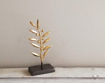 May include: A gold metal sculpture of an olive branch with small round details on the leaves. The sculpture is on a black rectangular base.