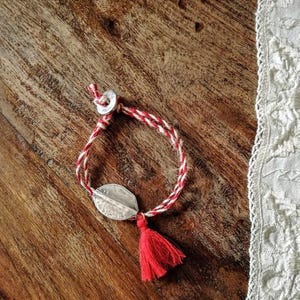 Op de afbeelding: Een rood en wit touw armband met een zilveren bedel en een rode kwast.