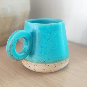 Puede incluir: Una pequeña taza de cerámica turquesa con un asa redondeada. La taza tiene una textura moteada y una base natural sin esmaltar. La taza está diseñada para bebidas calientes.