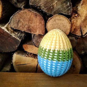 Op de afbeelding: Een gebreid eivormig ornament met een geel, groen en blauw gestreept patroon. Het ornament ligt op een stapel houtblokken.