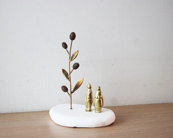 May include: A gold metal olive branch sculpture with two gold figurines standing on a white stone base.