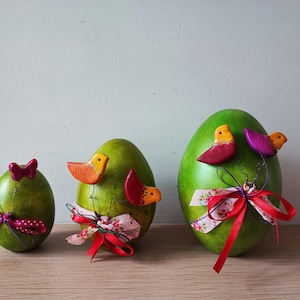 May include: Three green, egg-shaped decorative objects with wooden bird accents and colourful ribbons. The birds are red, orange, and purple. The ribbons are red and pink with floral patterns. The objects are arranged on a wooden surface.