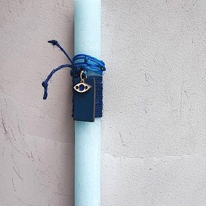 Può includere: Una candela azzurra con una corda blu e un ciondolo rettangolare blu scuro. Il ciondolo ha una cornice color oro e un design a occhio blu. La candela è appesa a uno spago bianco.