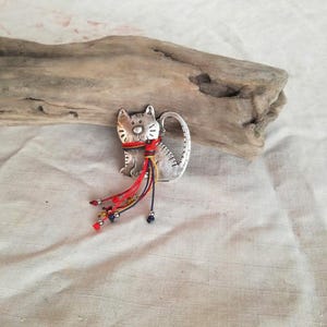 Pode incluir: Um amuleto de gato de metal prateado com uma echarpe vermelha, amarela e azul. O amuleto está sobre um fundo de tecido branco.