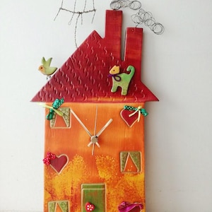 Puede incluir: Un reloj hecho a mano con forma de casa con un techo rojo, paredes naranjas y detalles verdes. El reloj tiene un pájaro, un gato y corazones en la parte delantera.