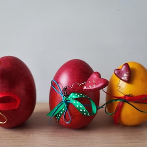 Op de afbeelding: Drie gedecoreerde paaseieren. Eén is rood met een groen-blauw lint en een rood hart. Een ander is rood met een rood lint en een oranje vlinder. De derde is geel met een rood lint en een rood hart.