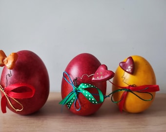 May include: Three decorated Easter eggs. One is red with a green and blue ribbon and a red heart. Another is red with a red ribbon and an orange butterfly. The third is yellow with a red ribbon and a red heart.