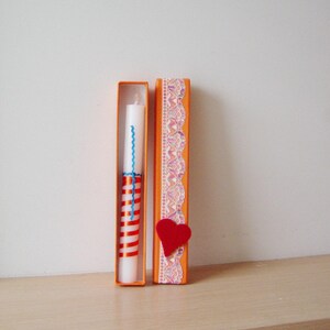 May include: A white candle with red and blue stripes is in an orange box with a heart-shaped design. The box is decorated with a white lace pattern.