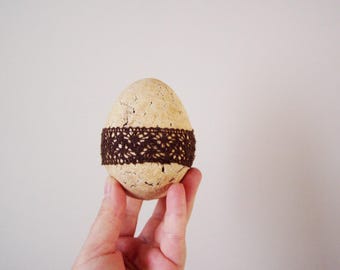 May include: A light brown egg with a brown lace ribbon wrapped around it. The egg is being held in a hand.
