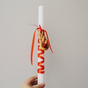 May include: A white candle with a red ribbon wrapped around it. A small, braided bread with a red berry is attached to the ribbon.