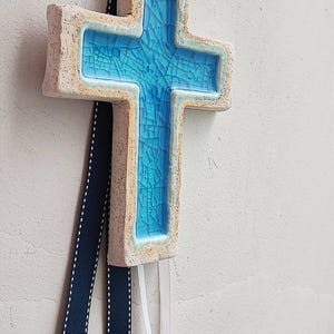 Könnte beinhalten: Ein Keramik-Kreuz mit hellbeigem Äußeren und einem leuchtend blauen, craquelé-Glas-Inneren. Oben ist ein marineblaues und weißes Band befestigt, darunter hängen weiße Bänder. Das Kreuz hängt an einer strukturierten, cremefarbenen Wand.