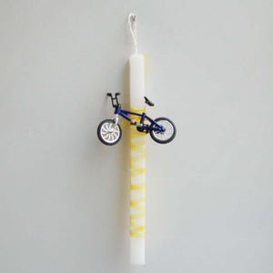 Puede incluir: Una vela de Pascua blanca con rayas amarillas y una pequeña bicicleta azul unida al lado.
