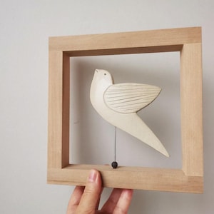 May include: A white ceramic bird sculpture with a wooden frame. The bird is suspended from the top of the frame by a thin wire.