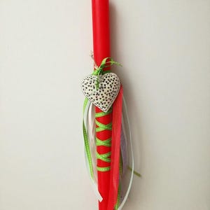 May include: A red Easter candle with a white heart-shaped ornament and green, white, and pink ribbons.