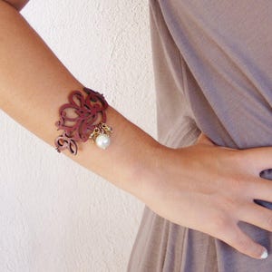 May include: A red leather bracelet with a delicate floral design and a pearl charm.