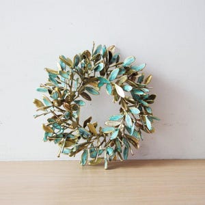 May include: A metal wreath with a circular design of leaves in gold and teal hues. The wreath is displayed on a wooden surface against a white wall. The leaves have a textured finish, adding detail to the decorative piece.