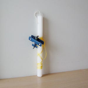 Può includere: Una candela bianca con una decorazione di aereo blu e un nastro giallo. La candela è legata con un nastro giallo e ha un piccolo aereo blu attaccato ad essa.