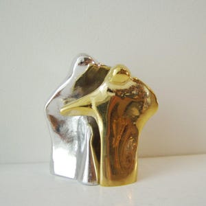 Puede incluir: Escultura abstracta de dos figuras, una plateada y otra dorada, abrazándose. Las figuras son estilizadas y tienen un diseño moderno.