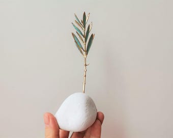 May include: A white ceramic vase in the shape of a rounded pebble with a gold-painted branch with teal leaves extending upwards.