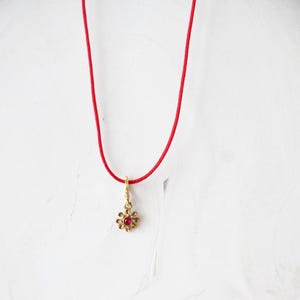 May include: A red string necklace with a gold flower pendant featuring a red gemstone in the center.