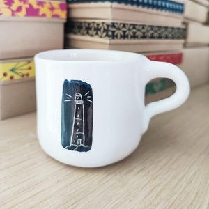 Op de afbeelding: Witte keramische mok met een zwart-witte illustratie van een vuurtoren met een lichtstraal die van boven schijnt. De mok heeft een afgeronde handgreep.