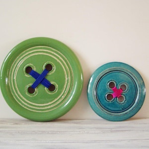 Puede incluir: Dos botones grandes de cerámica, uno verde con hilo azul y otro azul verdoso con hilo rosa.