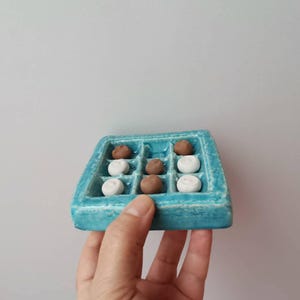 Può includere: Un gioco del tris in ceramica turchese con nove scomparti quadrati. I pezzi del gioco sono un mix di marrone e bianco, disposti nella griglia della tavola. La tavola è tenuta da una mano su uno sfondo semplice.