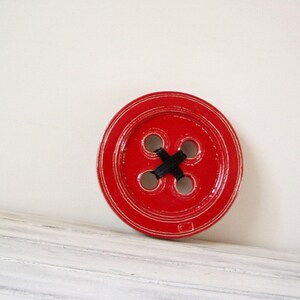 May include: A large, red wooden button with four black holes and a black X in the centre. The button has a raised, textured edge.