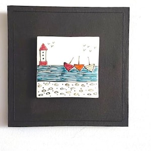 Puede incluir: Una obra de arte enmarcada que representa una escena costera. La imagen muestra un faro rojo y blanco, tres botes en rojo, naranja y blanco, y una costa con pequeños guijarros. La obra de arte está sobre un fondo negro.