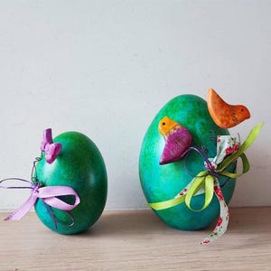 Könnte beinhalten: Zwei grüne Ostereier, die mit bunten Vögeln und Bändern verziert sind. Ein Ei hat einen rosa Vogel und eine lila Schleife, das andere einen orangefarbenen Vogel und eine grüne Schleife.