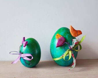 May include: Two green Easter eggs decorated with colorful birds and ribbons. One egg has a pink bird and a purple bow, the other has an orange bird and a green bow.