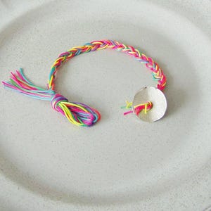 May include: A multi-colored braided bracelet with a silver charm. The bracelet is made of pink, yellow, blue, and green thread.