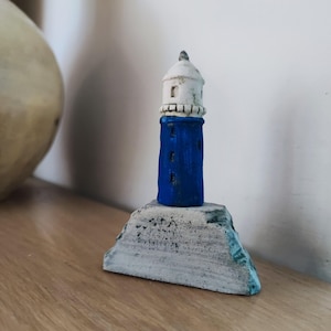 May include: A small blue and white ceramic lighthouse figurine on a white rock base. The lighthouse has a white roof and blue body with white windows.