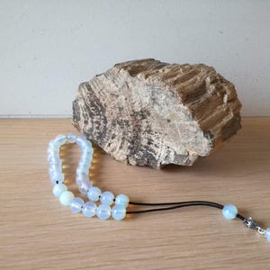 Puede incluir: Un collar de cuentas de ópalo blanco con un dije de plata. El collar está sobre una superficie de madera junto a una pieza de madera petrificada.