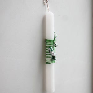 May include: A white candle with a green ribbon and a soccer shoe charm. The candle is hanging from a hook on a white wall.