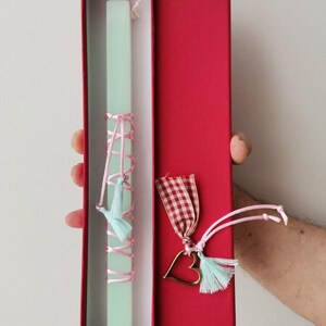 May include: A light green Easter candle with pink ribbon and blue tassels, in a red box with a heart-shaped charm and a red and white checkered ribbon.