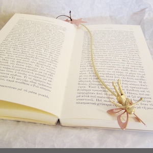 May include: A gold and copper butterfly bookmark with a long, thin, gold chain. The bookmark is resting on an open book with Greek text.