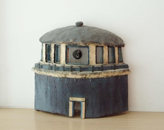 May include: A small, gray, round building with a thatched roof and a white door. The building has several windows with blue trim. There is a circular object in the window.