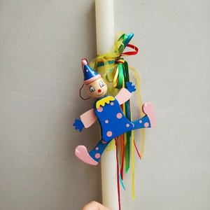 Puede incluir: Una vela de Pascua blanca con una muñeca de payaso de madera unida. El payaso lleva un traje de lunares azules y rosas y un sombrero azul. La vela está decorada con cintas de colores.