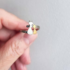 Op de afbeelding: Een zilverkleurige ring met twee kattenfiguren. Eén kat is zilverkleurig, de andere goudkleurig. De ring wordt tegen een lichte achtergrond gehouden. Een uniek sieraad.