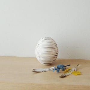 Op de afbeelding: Een witte, eivormige vaas van keramiek met bruine strepen. De vaas staat op een licht houten oppervlak met een paar kleine blauwe en gele bloemen.