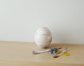 May include: A white ceramic egg-shaped vase with brown stripes. The vase is sitting on a light wood surface with a few small blue and yellow flowers.