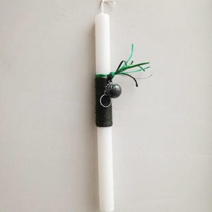 May include: A white candle with a black and green ribbon and a small black bell charm. The candle is decorated with a green and black ribbon and a small black bell charm.