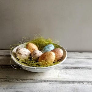 Peut inclure: Un bol blanc rempli d'herbe verte et de six œufs de Pâques tachetés. Les œufs sont dans des tons de bleu, vert et marron.