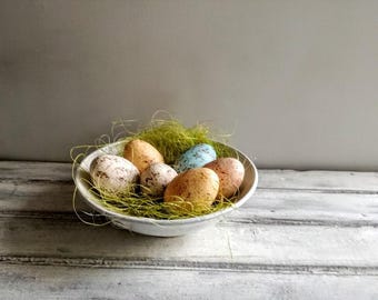 May include: A white bowl filled with green grass and six speckled Easter eggs. The eggs are in shades of blue, green, and brown.