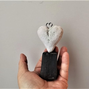 Op de afbeelding: Een wit hartvormig beeldje met een draadlus bovenop, dat op een zwarte houten voet staat.