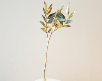 May include: A gold metal sculpture of an olive branch with green leaves, standing on a white stone base.