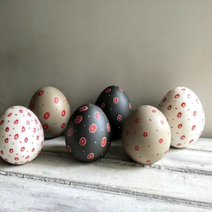 Peut inclure: Six œufs de Pâques décoratifs avec un motif tacheté. Les œufs sont peints dans diverses couleurs, dont le blanc, le beige et le gris foncé, chacun orné de pois rouges. Ils sont disposés sur une surface en bois vieilli peint en blanc.